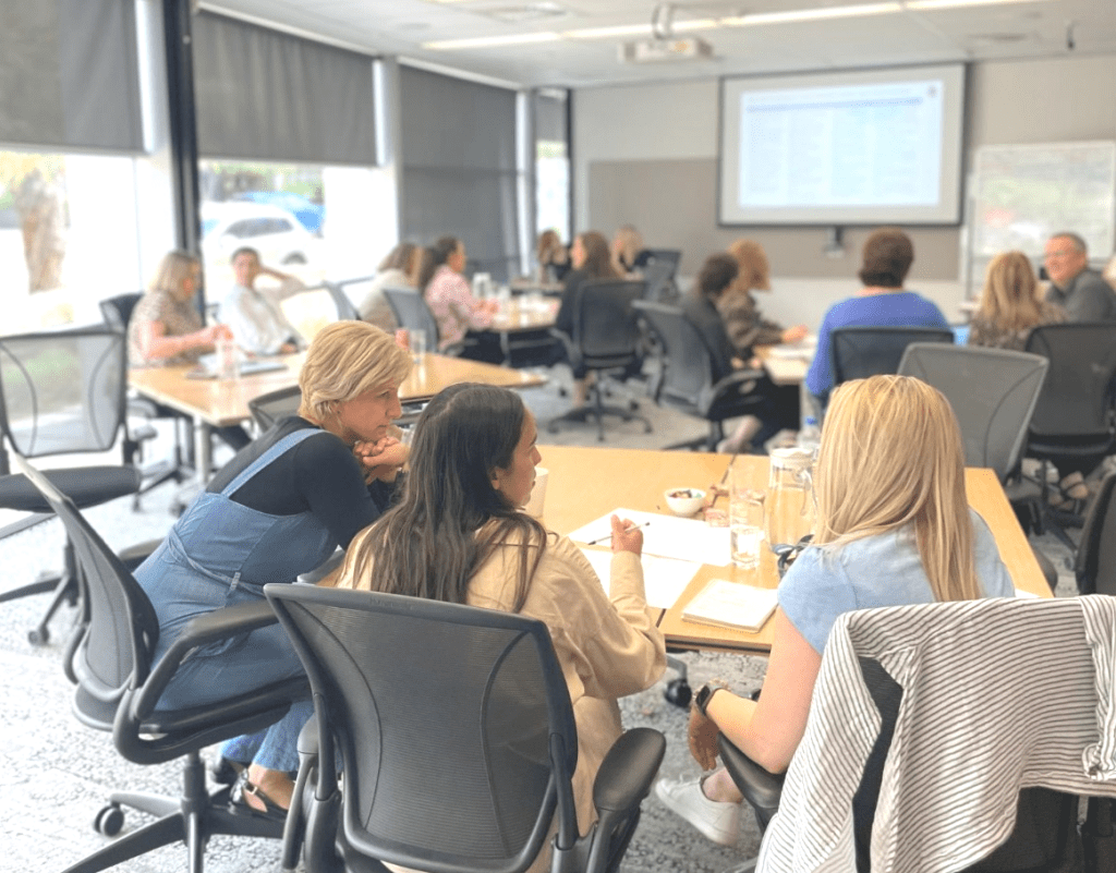 Image depicting a group of MercyCare staff sitting in a large office style room situated around four tables. All people are in discussion with each other. At the front of the room is a presentation on a screen and a whiteboard which are blurred.