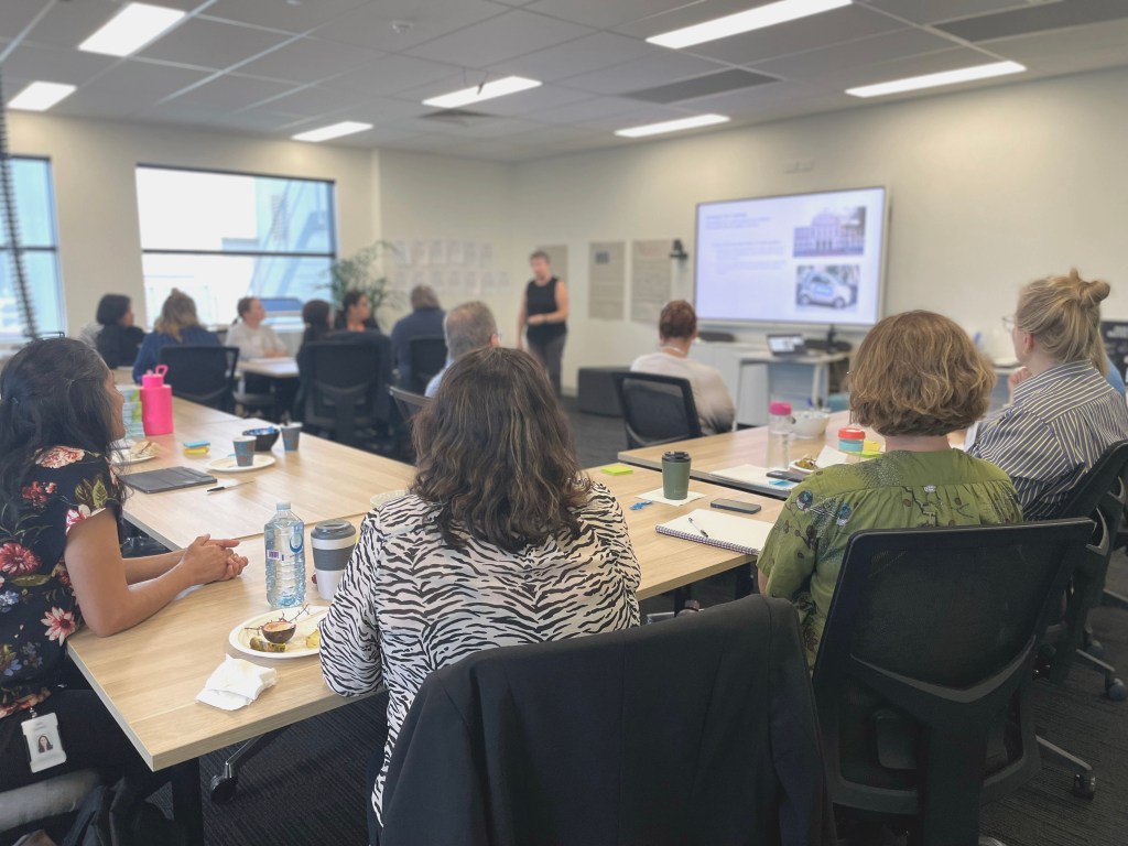Image showing staff from IPC Health sitting at a large table U-Shape looking towards the front of the room where Cathy Balding from Qualityworks gives a presentation